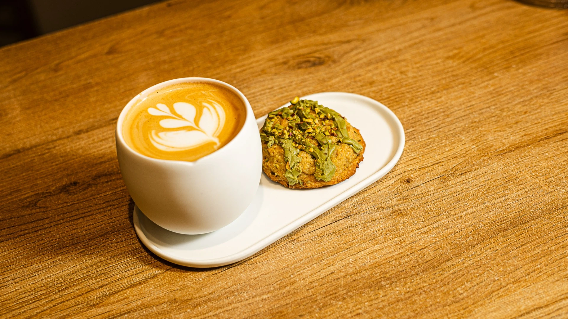 Taza de café con arte latte y galleta con pistacho en bandeja blanca sobre mesa de madera, ideal para tienda de café de especialidad.