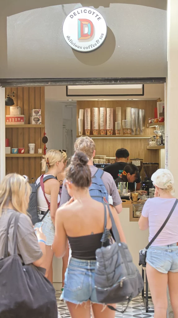 Clientes haciendo fila en mostrador de tienda de café de especialidad Delicotte con decoración de madera y vasos apilados.