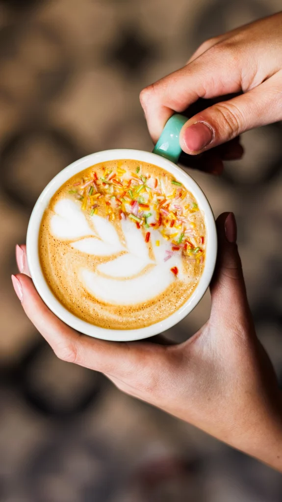 Taza de café con arte latte decorado con chispas de colores, sostenida por dos manos en la tienda de café de especialidad.