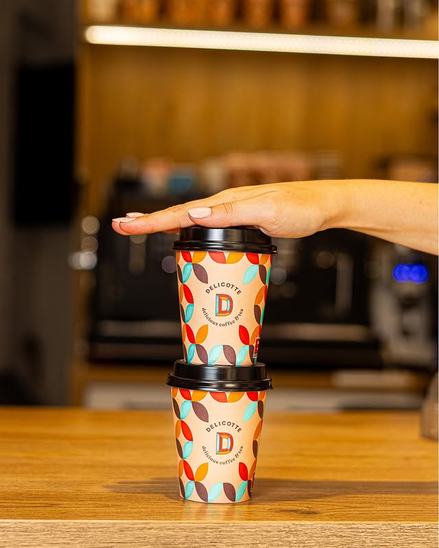 Dos vasos de café para llevar con diseño colorido apilados y una mano sobre el vaso superior en tienda de café de especialidad.