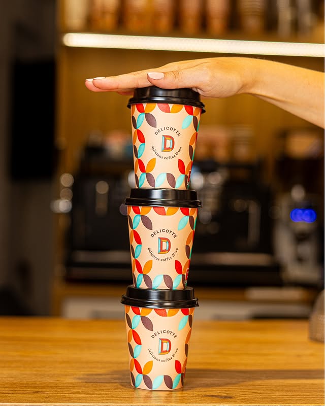 Tres vasos de café para llevar con diseño colorido apilados y una mano sobre el vaso superior en tienda de café de especialidad.