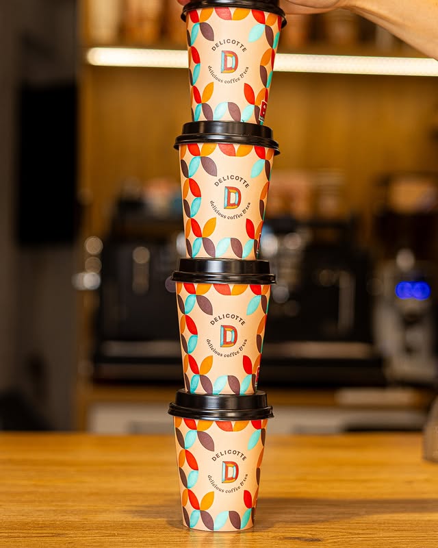 Cuatro vasos de café para llevar con diseño colorido apilados en torre y una mano sujetando el vaso superior en tienda de café de especialidad.