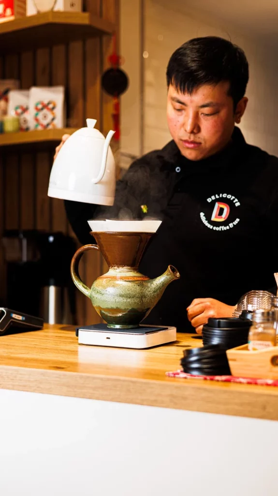 Barista de tienda de café de especialidad preparando café filtrado con tetera y gotero de cerámica sobre balanza digital.