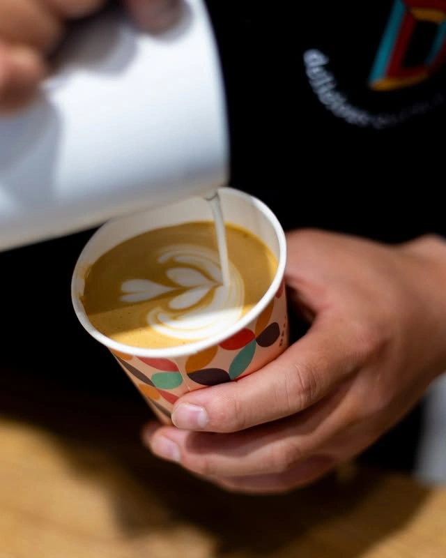 Barista sirviendo arte latte en vaso para llevar con diseño colorido en tienda de café de especialidad.