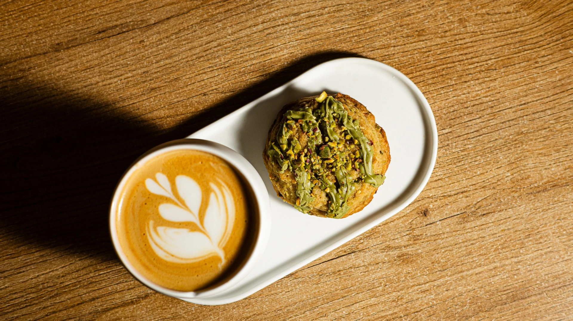 Taza de café con arte latte y galleta con pistacho en plato blanco sobre mesa de madera, ideal para tienda de café de especialidad.